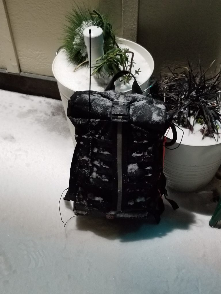 Snow and ice covered backpack set on a snow-covered sidewalk with counterpoise extending down and antenna extending up.