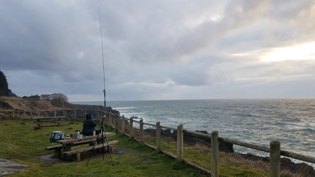 An antenna is mounted on a tripod in the foreground. Directly behind it is a park bench with someone sitting on it and some radio equipment with a cooler. The background is a fenced-in green area and the Pacific ocean and steep rocks in the background.