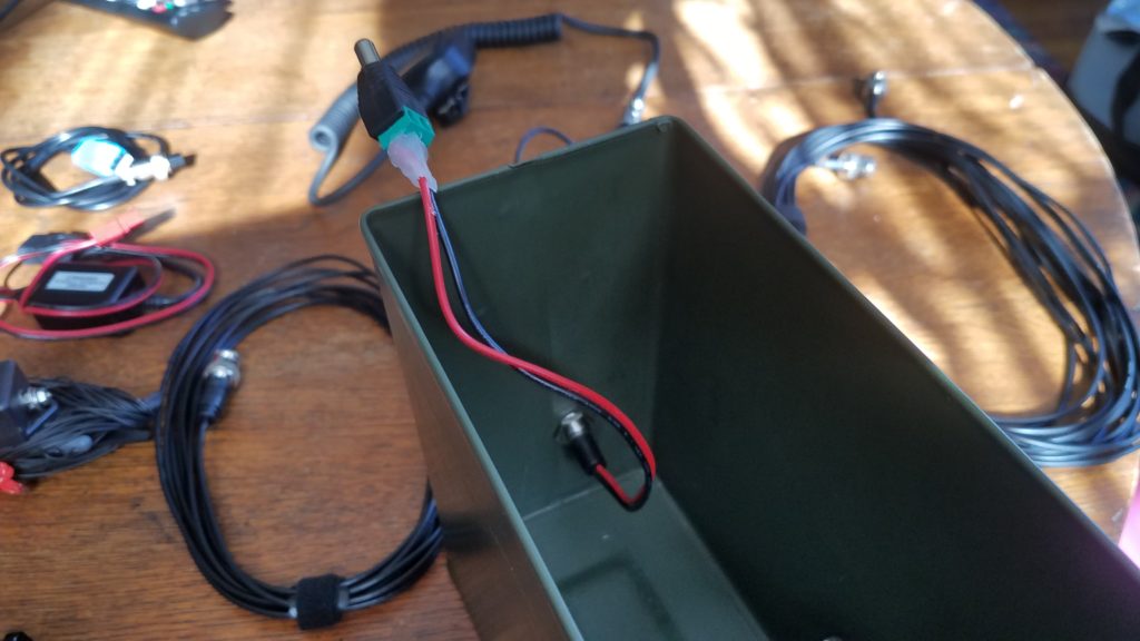 Inside view of an ammo can with barrel connector jack mounted on the front and the DC barrel connector connected to the wire coming from the jack. The barrel connector end's wires are hot glued to the barrel connector. The ammo can is sitting on a wood table and some wires and connectors are sitting on the table in the background.