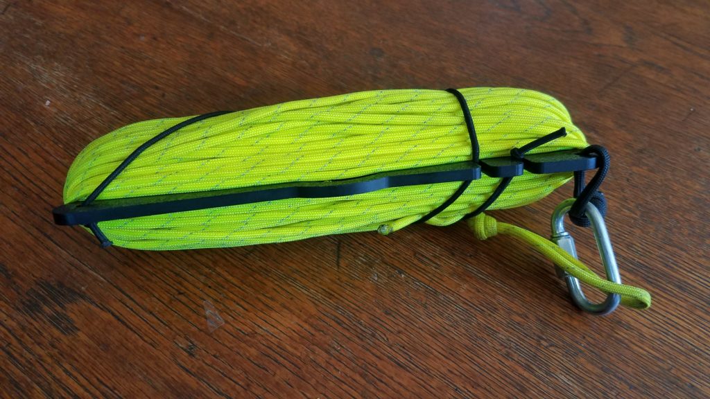 Chameleon wire winder with bright yellow paracord wrapped around it and secured to the wire winder by an elastic band. The paracord and wire winder are connected together with a quick link.