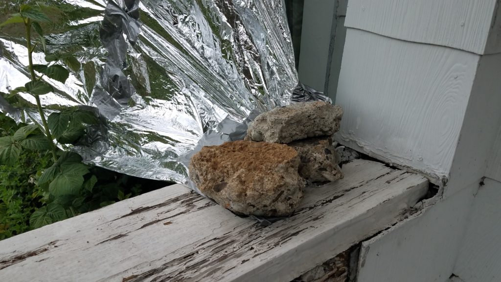 One corner of a mylar blanket being covered in duct tape and weighed down by three pieces of broken concrete sitting on a piece of wood in front of some siding.