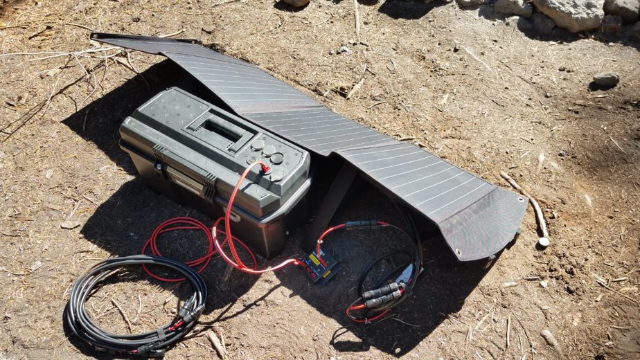 Power cables connecting a 100Ah battery box, solar charger, and solar panel sitting on the ground in the sun.