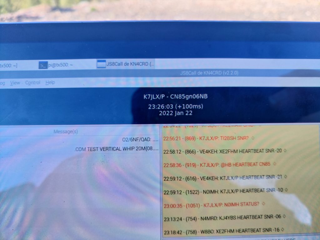 A photograph of the screen of a tablet showing the JS8Call application running. A callsign, timestamp, and 10-digit maidenhead coordinate are displayed prominently in the photo along with a screen showing contacts with other stations.