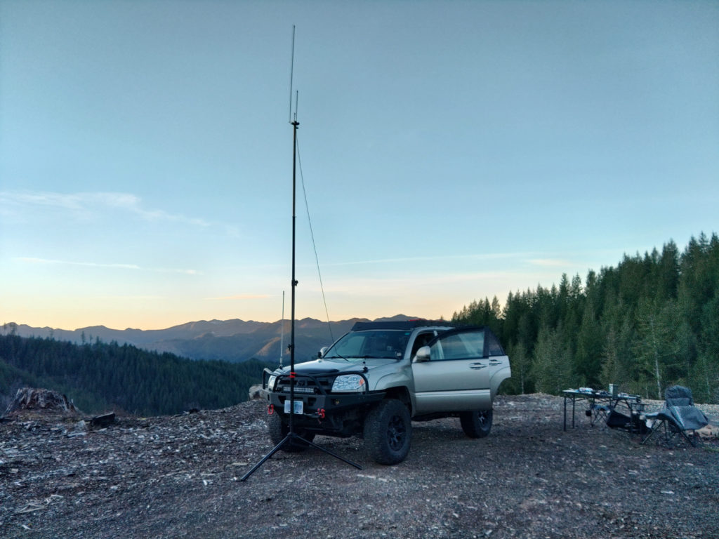A 4Runner with an antenna mast tied to the front bumper and connected to the vehicle with feedline. There's a camping table and chairs to one side and in the background are trees, a valley and a mountain on the other side of the valley.