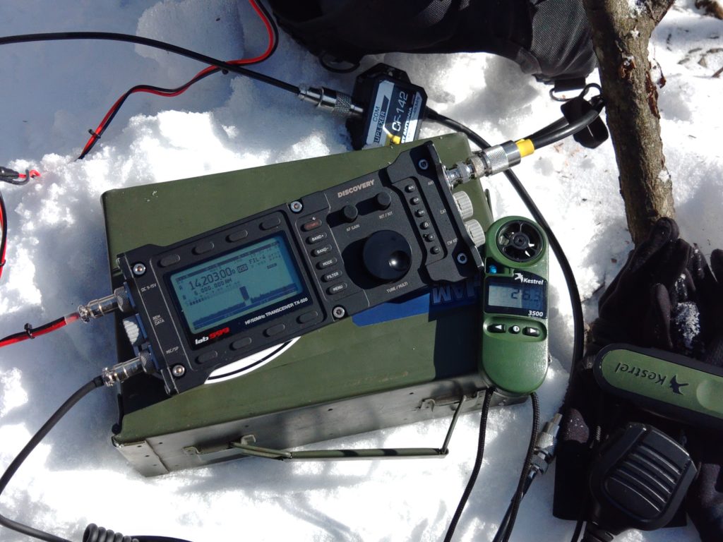 A Lab599 TX-500 radio sitting on top of an ammo can with a Kestrel portable weather station reading 26.3F. The radio is connected to a duplexer which is laying on the snow.