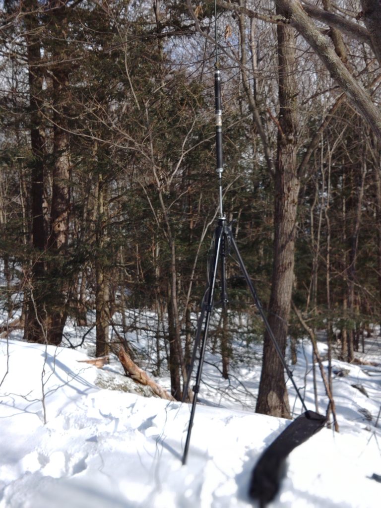 A Superantenna is set up with a 2m coil installed on a tripod. Trees and brush are visible in the background.