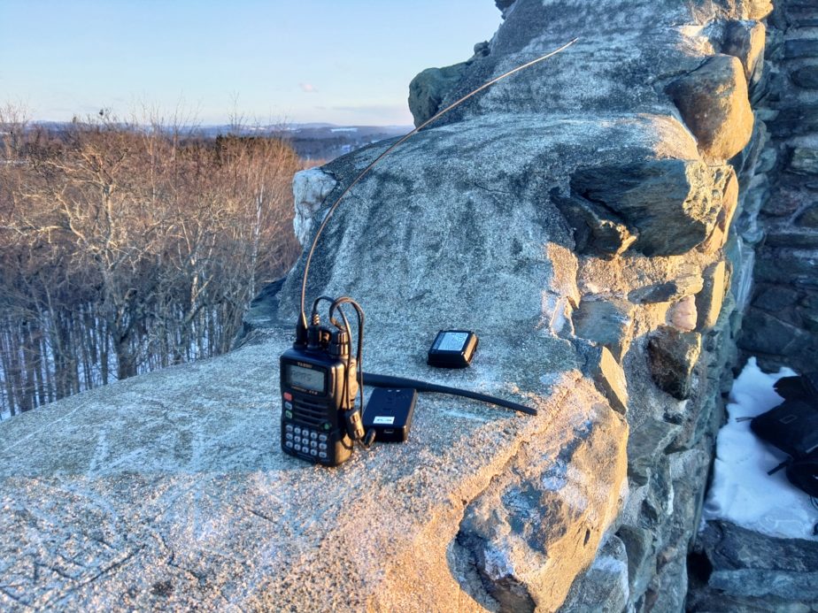 A handheld radio and associated components sits on a stone wall with trees and snow in the background. The antenna coming from the radio is bent at a 90 degree angle.